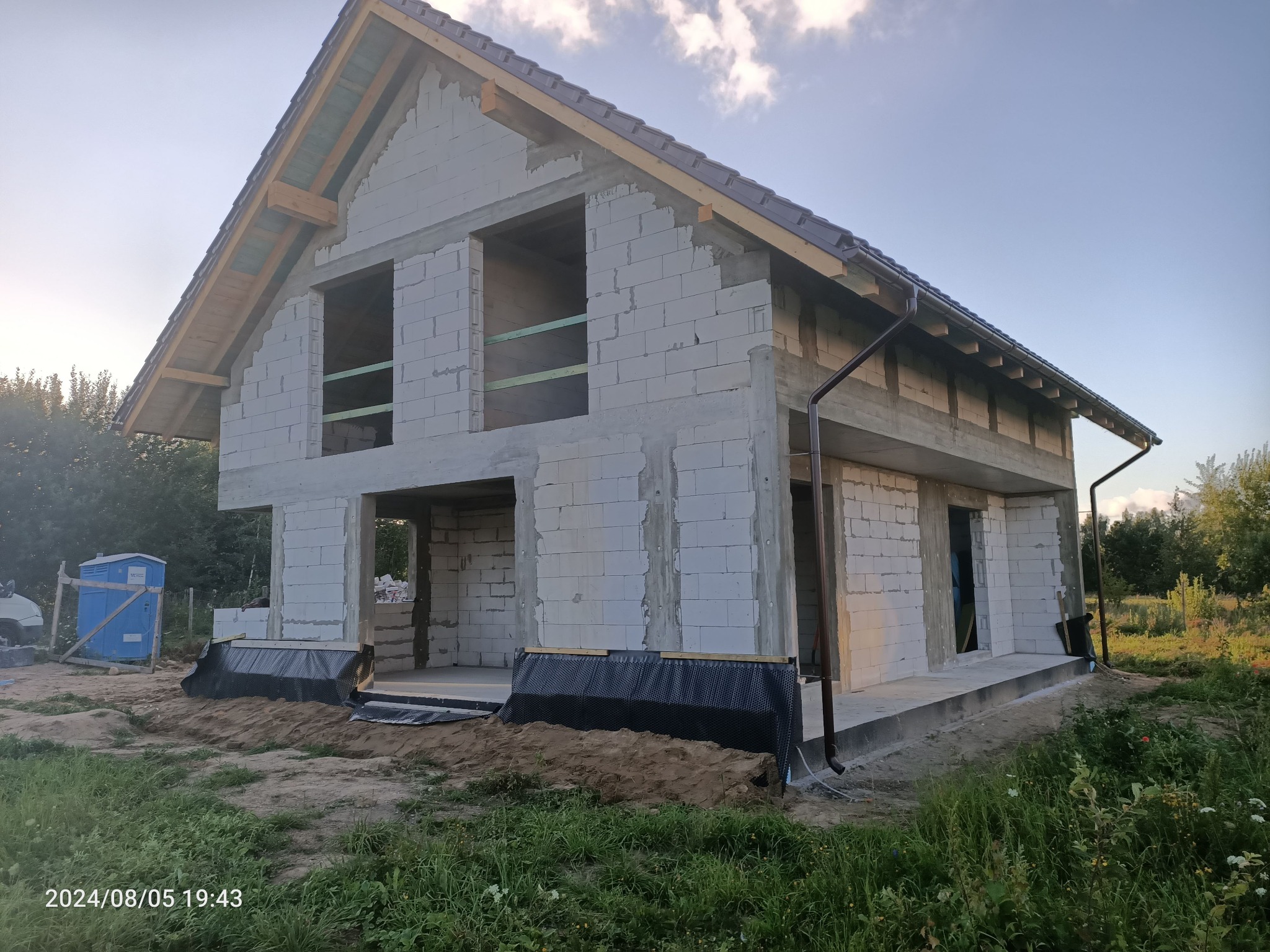 Surowy stan domu z białego bloczka, widoczne niezabezpieczone okna i drzwi, dach pokryty dachówką, rynny zamontowane, teren wokół domu częściowo obsypany ziemią, w tle widoczna przenośna toaleta.