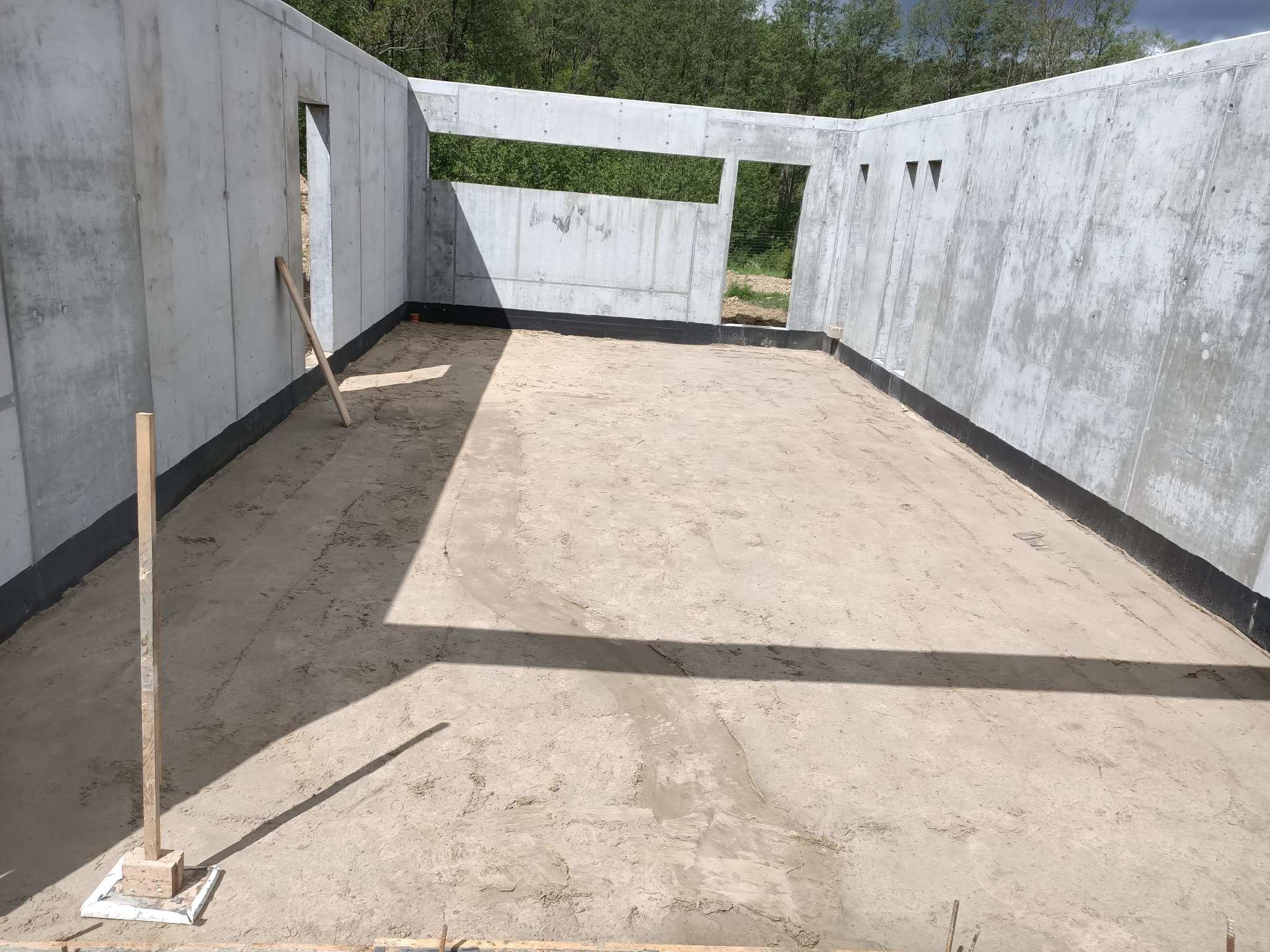 Surowy stan piwnicy z betonowymi ścianami, widoczne otwory okienne i drzwiowe, podłoże przygotowane pod wylewkę, drewniane podpory w narożnikach.