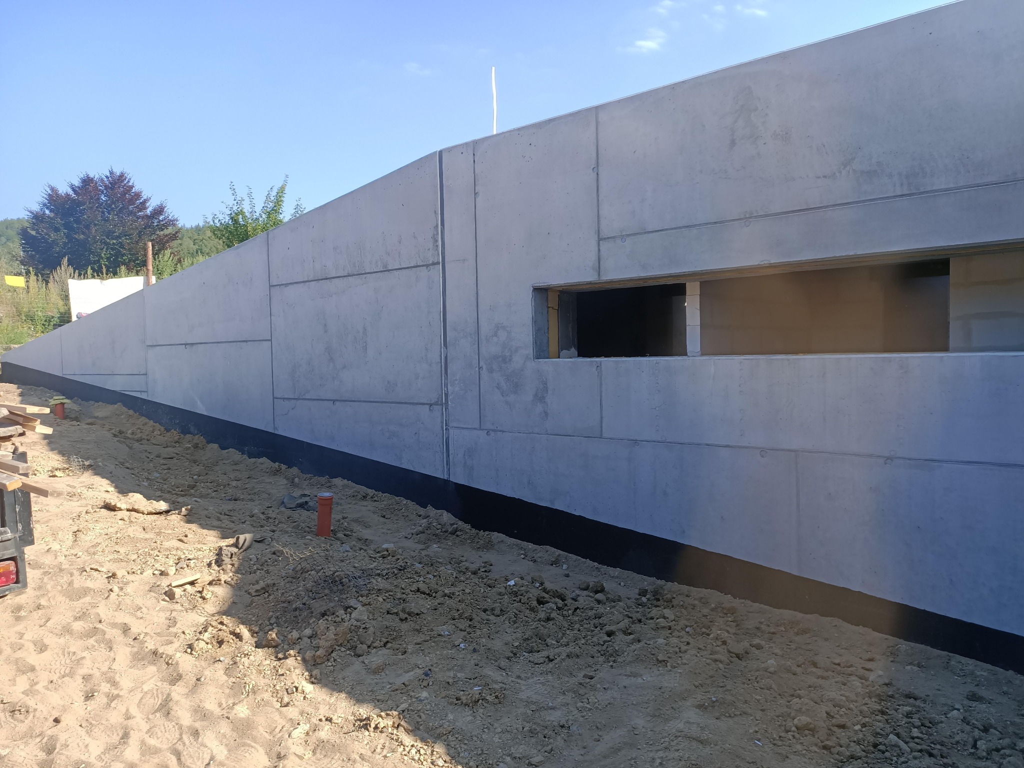 Surowy, betonowy fundament z otworem okiennym w trakcie budowy, z widoczną izolacją przeciwwilgociową i przygotowanym terenem wokół.