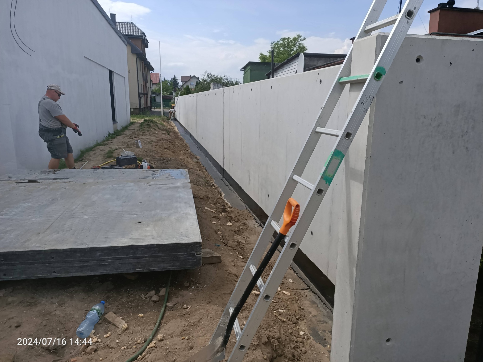 Wznoszenie betonowego ogrodzenia panelowego z widocznymi otworami technologicznymi, pracownik z narzędziami w tle, aluminiowa drabina oparta o mur, przygotowane panele na ziemi.