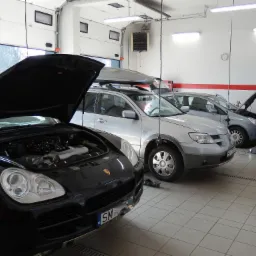 Częściowo widoczny czarny Porsche Cayenne z otwartą maską, stojący w jasnym warsztacie samochodowym z trzema innymi samochodami, w tle brama garażowa i oświetlenie sufitowe.