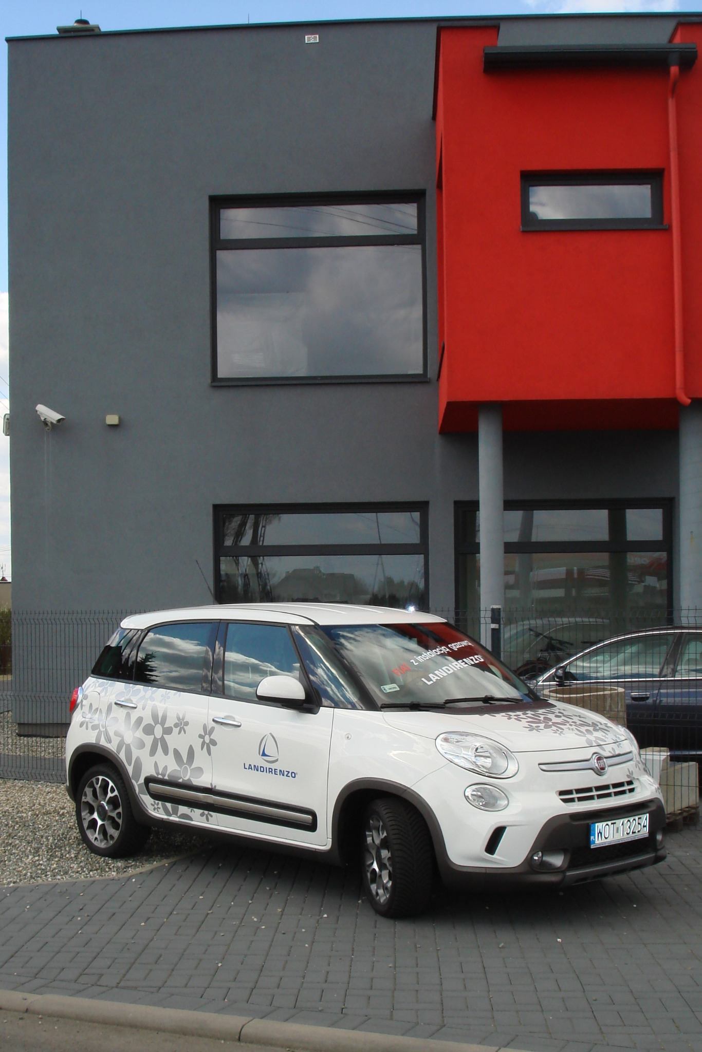 Biały samochód z logo Landirenzo, ozdobiony kwiatowymi wzorami, zaparkowany przed nowoczesnym budynkiem z szarą i czerwoną elewacją.