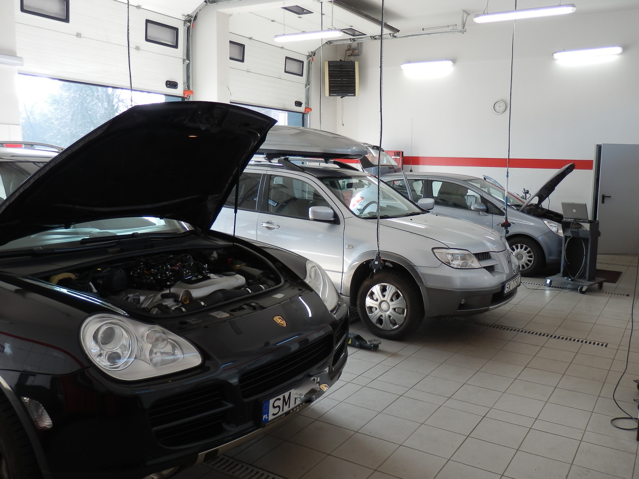 Częściowo widoczny czarny Porsche Cayenne z otwartą maską, stojący w jasnym warsztacie samochodowym z trzema innymi samochodami, w tle brama garażowa i oświetlenie sufitowe.