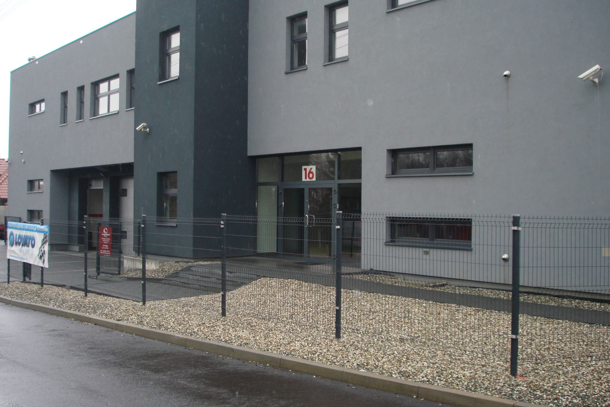 Szary budynek z numerem 16 nad wejściem, ogrodzony metalową siatką, z widocznym banerem reklamowym Lovato Auto Gaz.