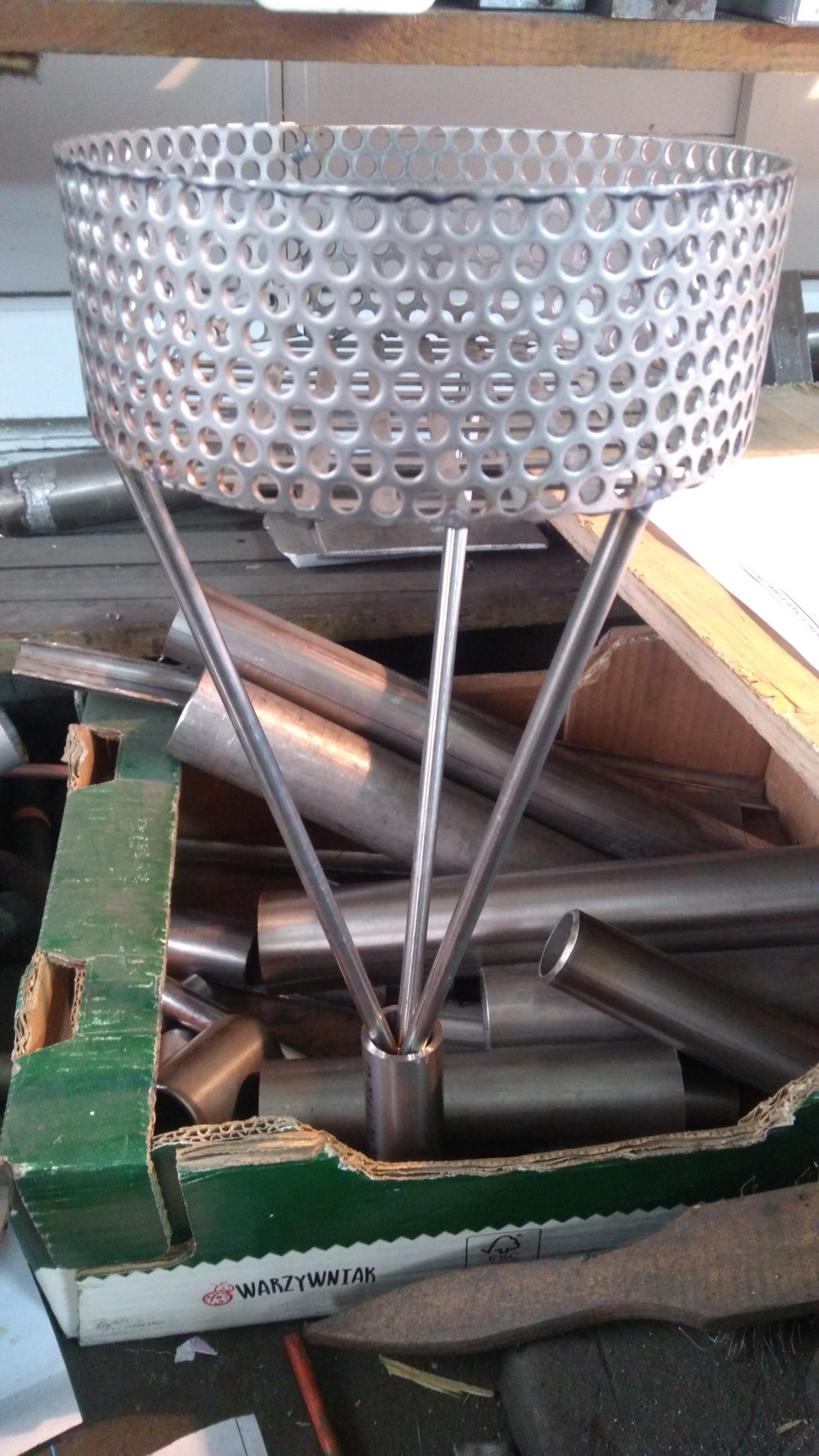 Metalowy kwietnik z perforowanym koszem i trzema nogami, wykonany w Pionkach, widoczny w otoczeniu metalowych rur i kartonowego pudła.