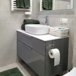 Szara szafka łazienkowa z umywalką nablatową, chromowaną baterią i białym blatem, uzupełniona o biały koszyk z akcesoriami, podgrzewany wieszak na ręczniki z ciemnozielonymi ręcznikami i lustro...