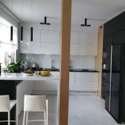 Nowoczesna biała kuchnia z wyspą i drewnianym panelem działowym, oświetlona wiszącymi lampami tubowymi i punktowymi, z owocami na blacie.