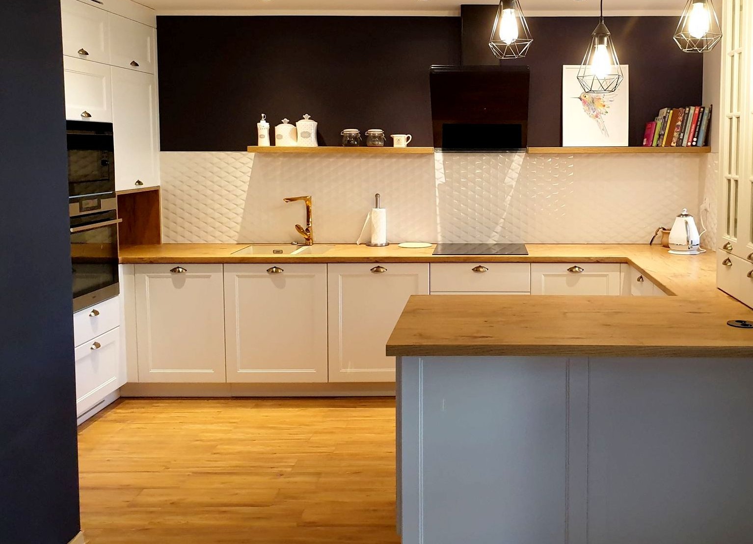 Jasna kuchnia z drewnianymi blatami, białymi szafkami i złotą armaturą, oświetlona wiszącymi lampami w geometrycznych kloszach, z widoczną wyspą kuchenną i drewnianą podłogą.