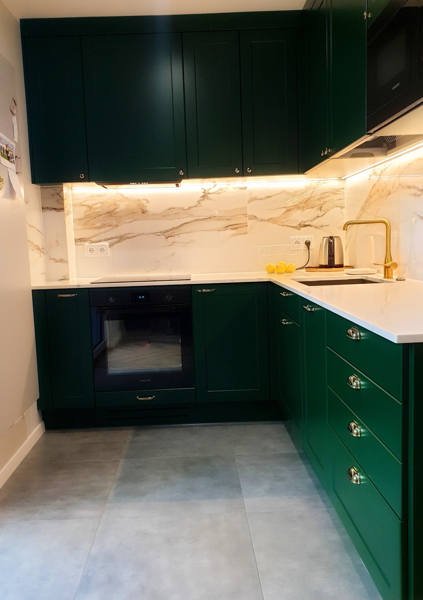 Nowoczesna kuchnia w odcieniach butelkowej zieleni z białym blatem i złotą armaturą, marmurowa ściana nad blatem z oświetleniem LED.