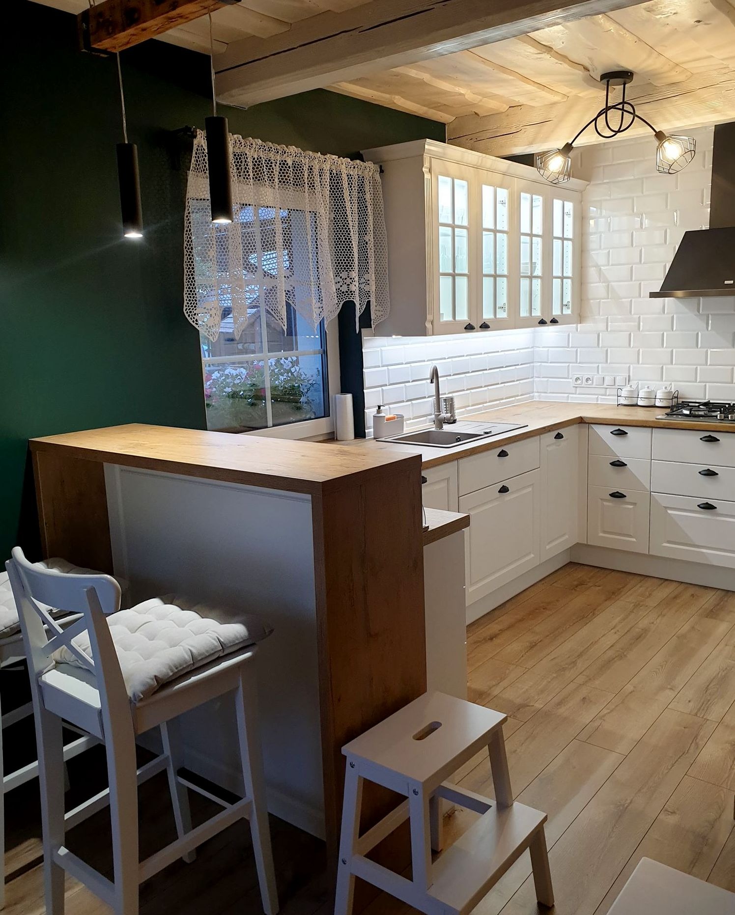 Jasna kuchnia z drewnianym blatem i białymi szafkami, oświetlona lampami wiszącymi i lampą sufitową, z drewnianym barkiem i krzesłami barowymi, oraz schodkami dla dzieci.