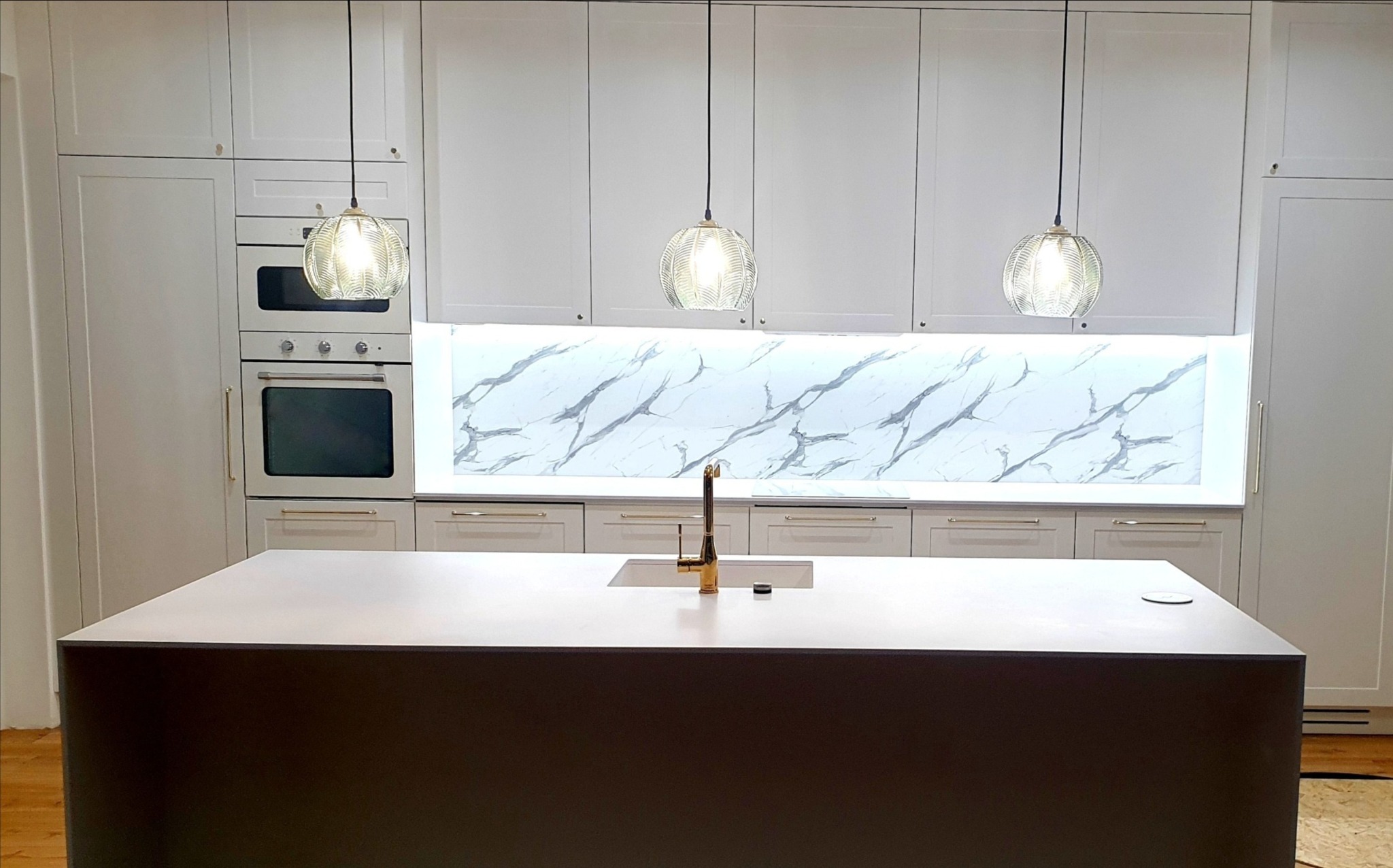 Nowoczesna kuchnia z wyspą, białe szafki z delikatnymi złotymi uchwytami, marmurowy panel nad blatem, złota bateria i trzy szklane lampy wiszące.