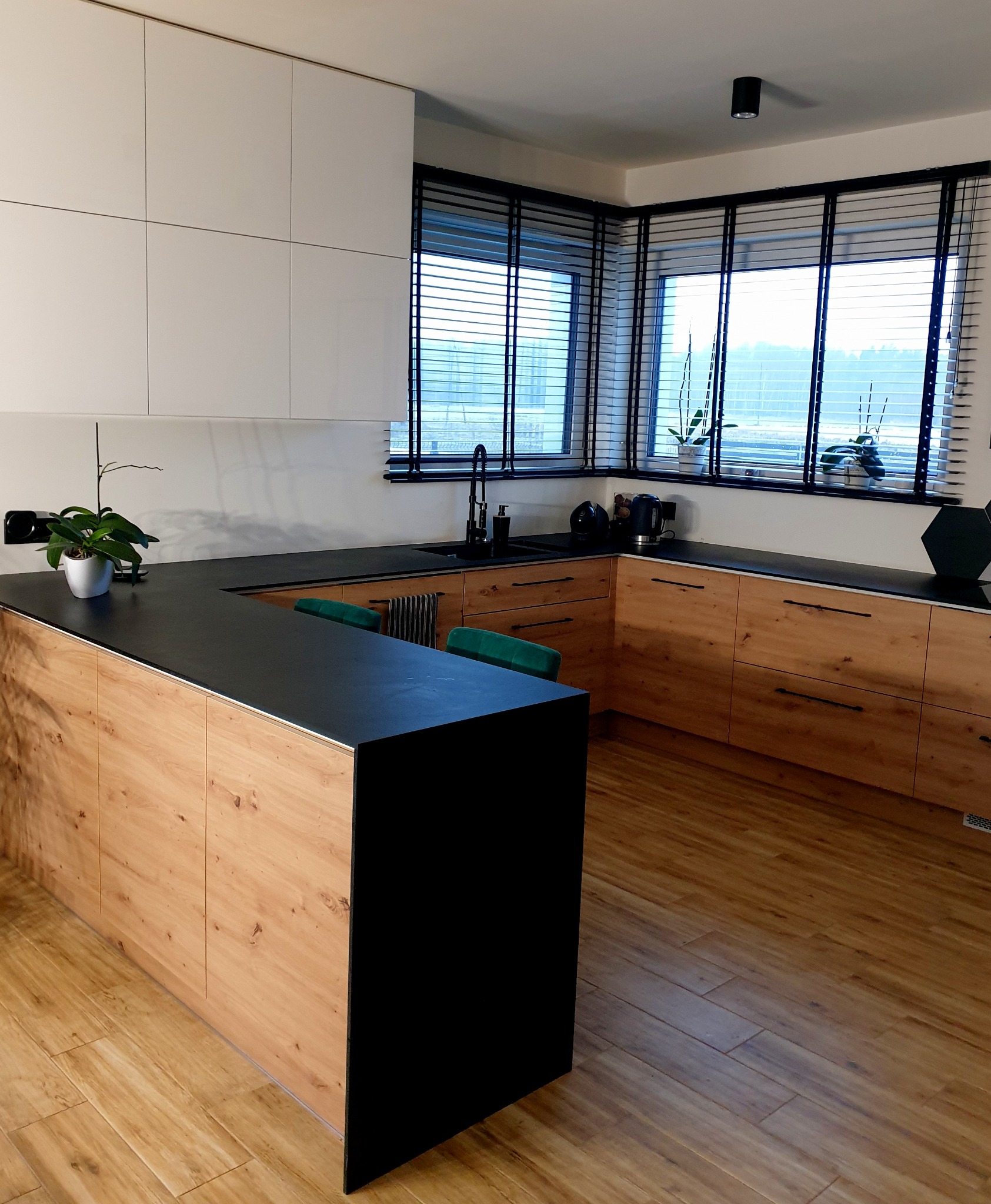 Nowoczesna kuchnia z drewnianymi frontami szafek, czarnym blatem i wyspą, oświetlona światłem dziennym wpadającym przez okno z żaluzjami.