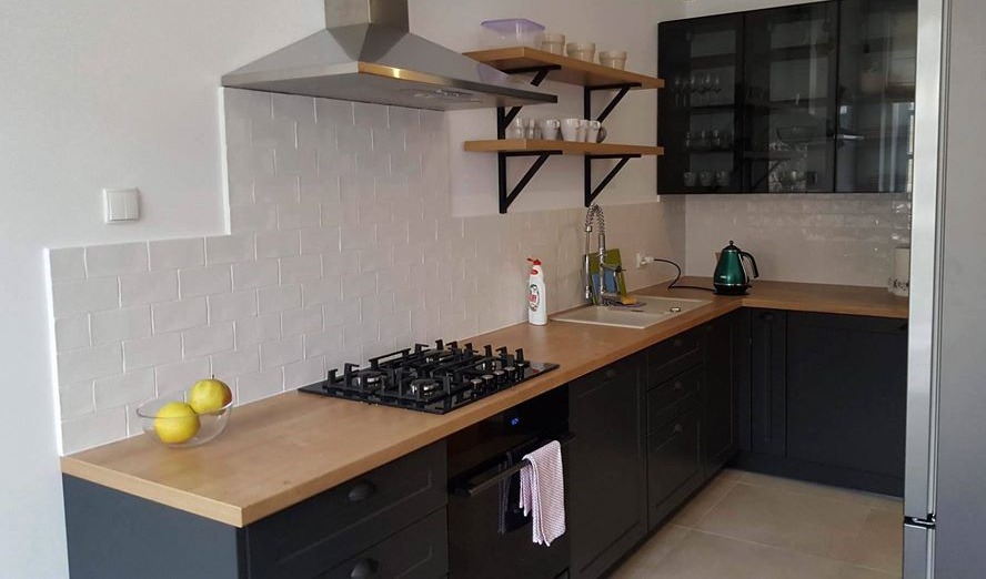 Nowoczesna kuchnia z ciemnymi szafkami, drewnianymi blatami, białymi kafelkami na ścianie i otwartymi półkami z czarnymi wspornikami, na blacie misa z jabłkami.