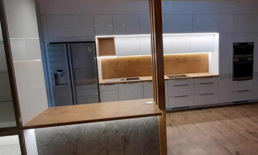 Nowoczesna kuchnia na wymiar z białymi, matowymi frontami, drewnianym blatem, podświetleniem LED i wyspą z marmurowym wykończeniem.