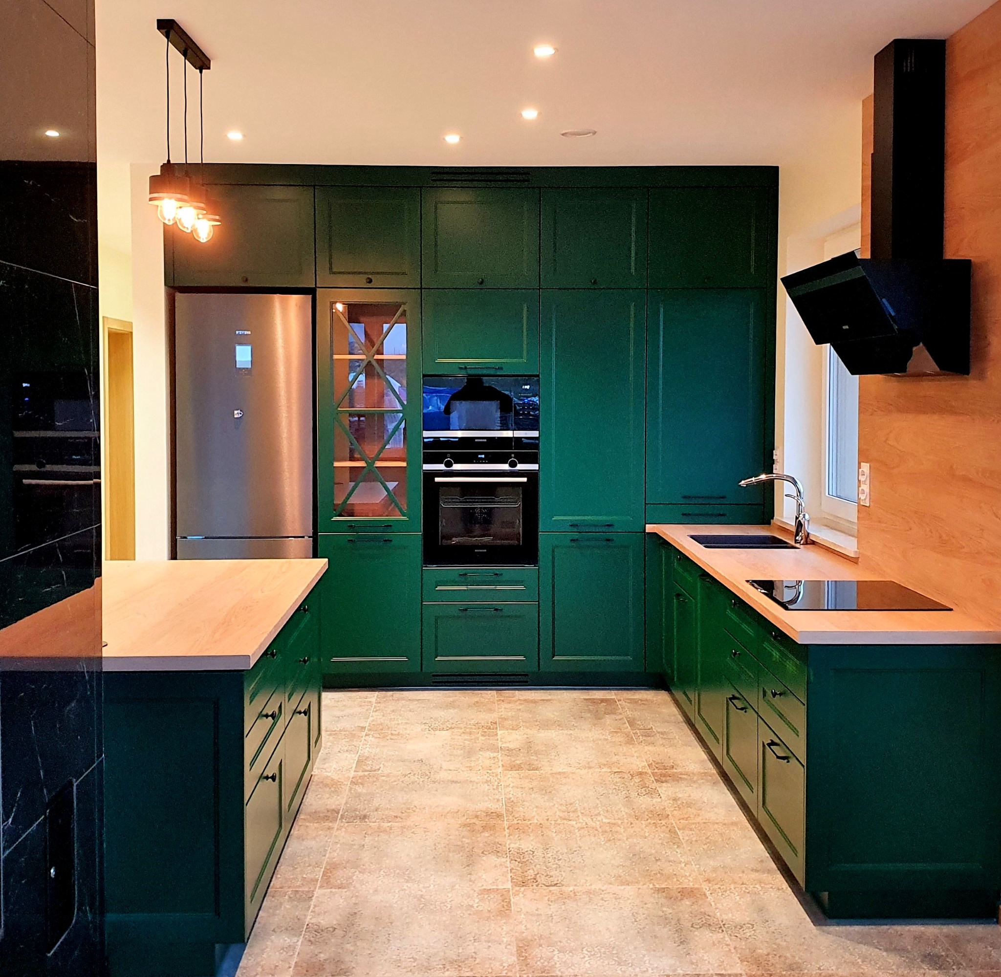 Nowoczesna kuchnia z ciemnozielonymi szafkami, drewnianymi blatami i wyspą z czarnym marmurem, oświetlona trzema wiszącymi lampami z żarówkami Edisona.