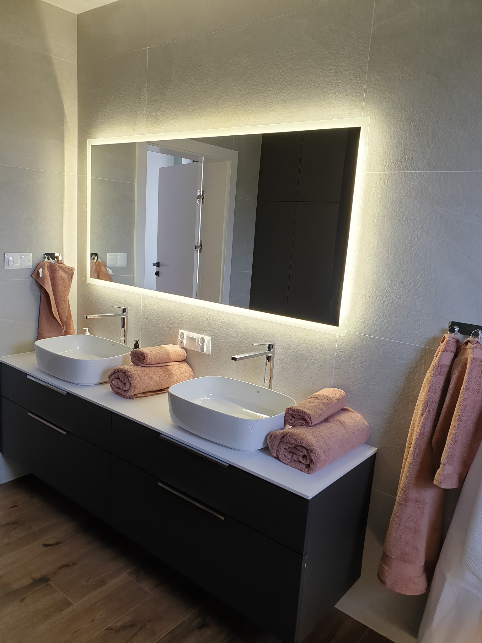 Łazienka z dwoma umywalkami nablatowymi, czarną szafką pod umywalki i podświetlanym lustrem, ręczniki w odcieniach różu, płytki imitujące kamień.