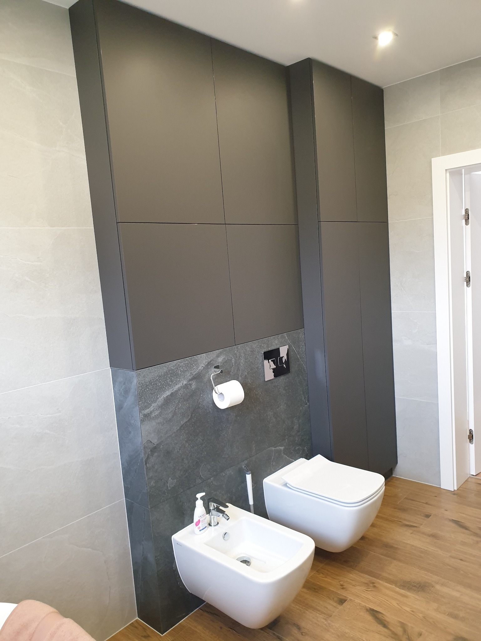Nowoczesna łazienka z zabudową szafkową w kolorze grafitowym, wiszącą toaletą i bidetem na tle płytek imitujących kamień.