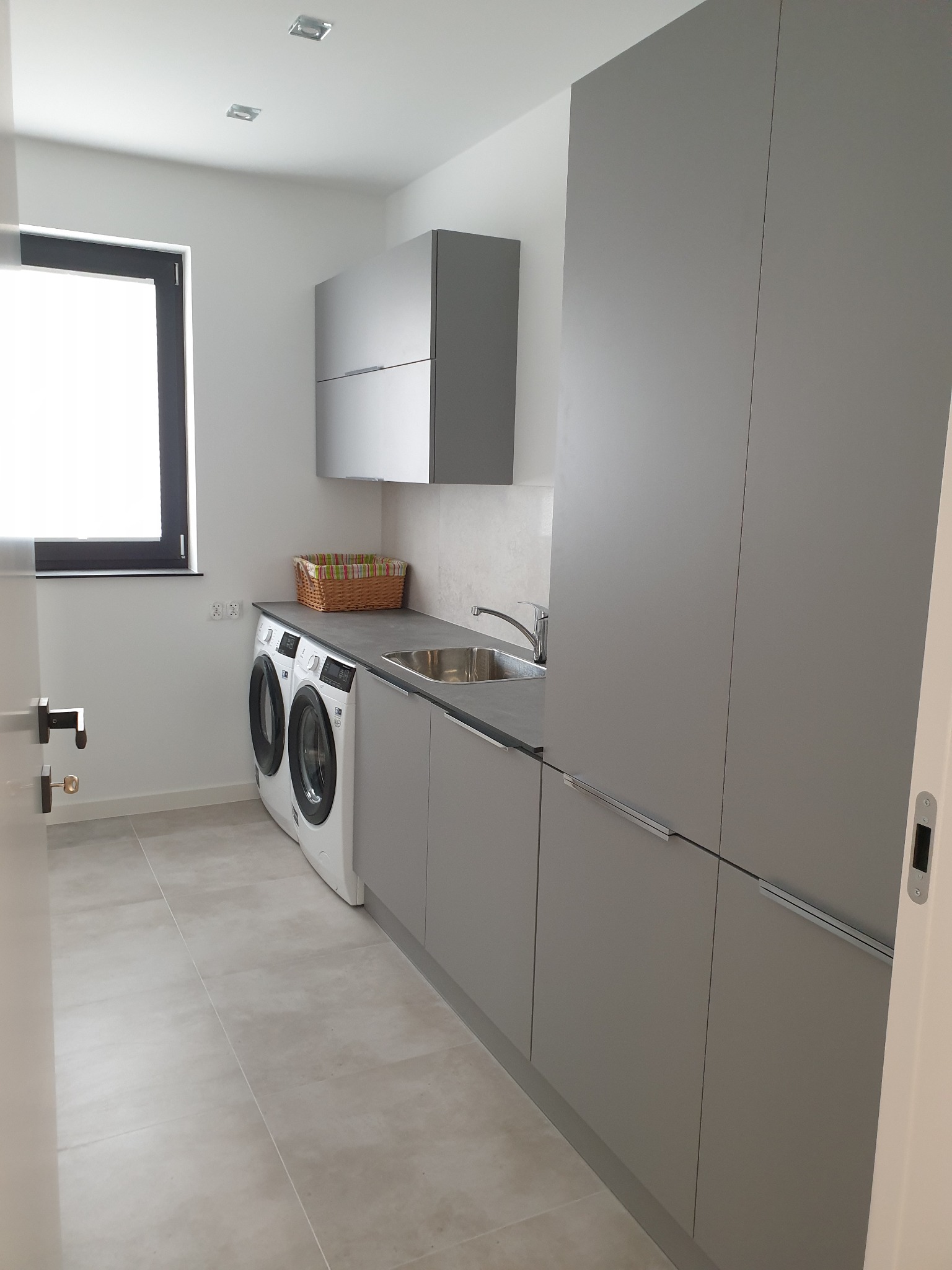 Nowoczesna pralnia w szarościach z dwiema pralkami, blatem, szafkami wiszącymi i wysoką zabudową, uzupełniona o wiklinowy koszyk na blacie.
