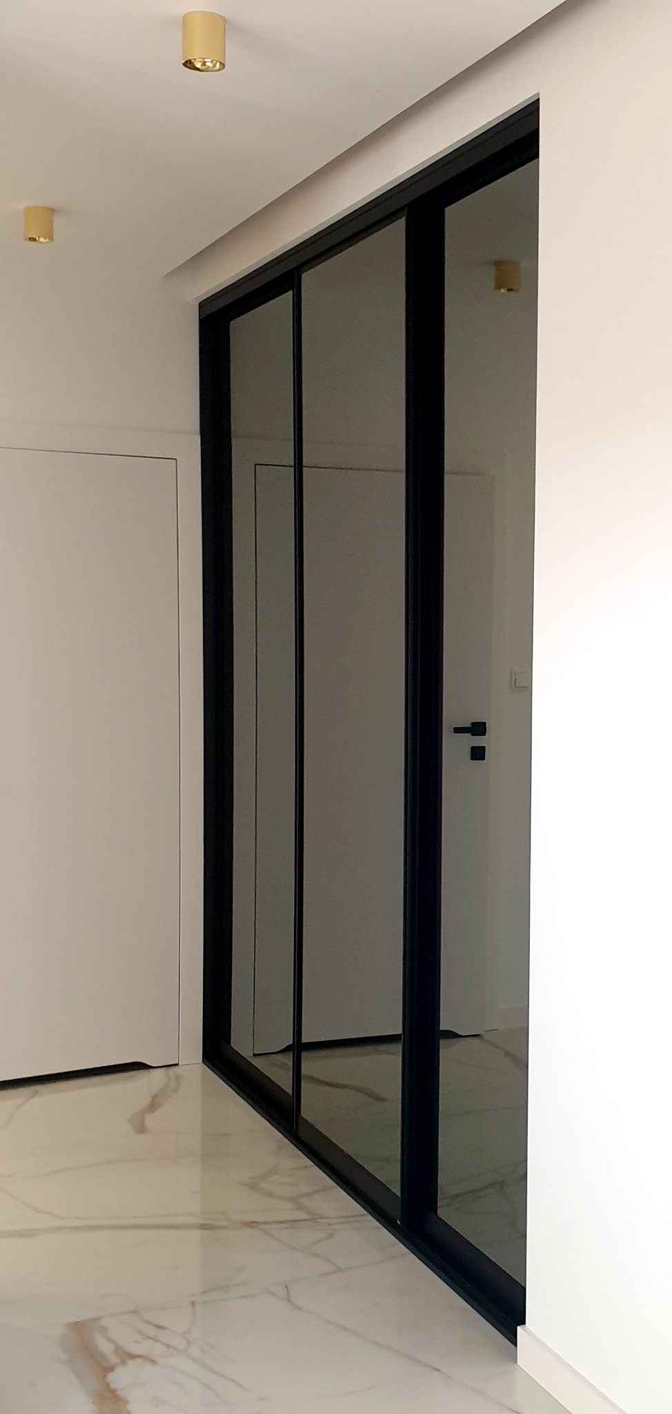 Nowoczesna szafa wnękowa z przesuwnymi, lustrzanymi drzwiami w czarnej ramie, widoczna z boku na tle białych ścian i marmurowej podłogi, oświetlona złotymi lampami sufitowymi.
