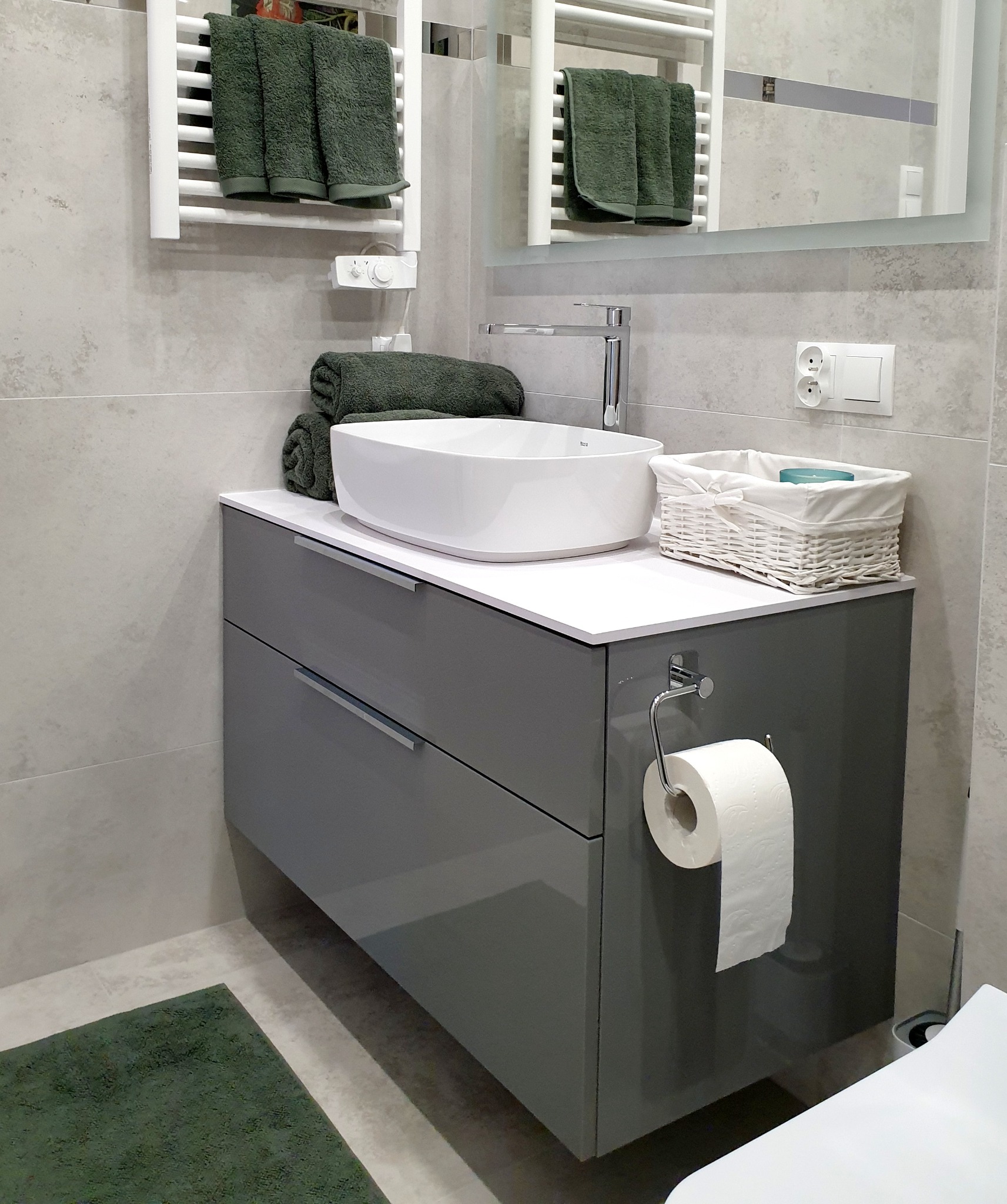 Szara szafka łazienkowa z umywalką nablatową, chromowaną baterią i białym blatem, uzupełniona o biały koszyk z akcesoriami, podgrzewany wieszak na ręczniki z ciemnozielonymi ręcznikami i lustro...