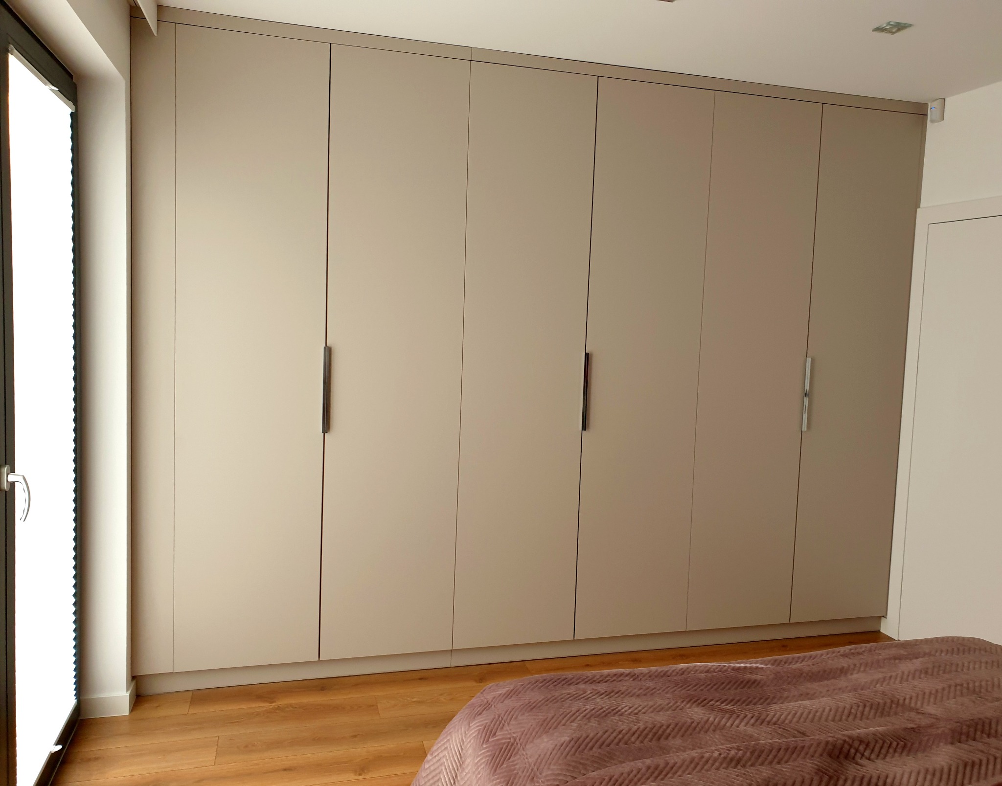 Beżowa szafa wnękowa z długimi, pionowymi uchwytami, widoczna częściowo z perspektywy łóżka z brązową narzutą i drewnianą podłogą.