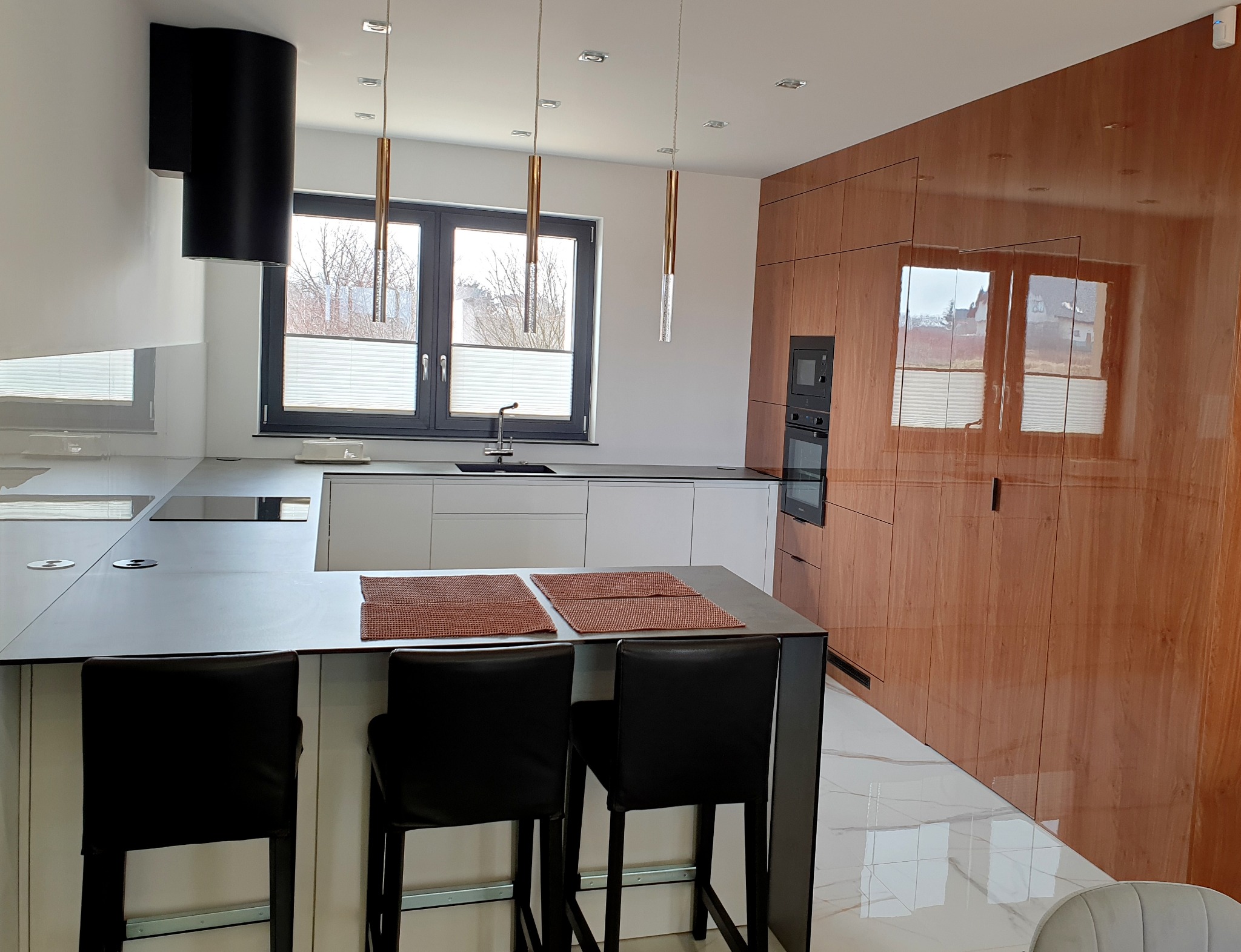 Nowoczesna kuchnia z białymi szafkami, ciemnoszarym blatem wyspy, drewnianą zabudową ścienną, czarnym okapem cylindrycznym i trzema wiszącymi lampami z elementami w kolorze miedzi.