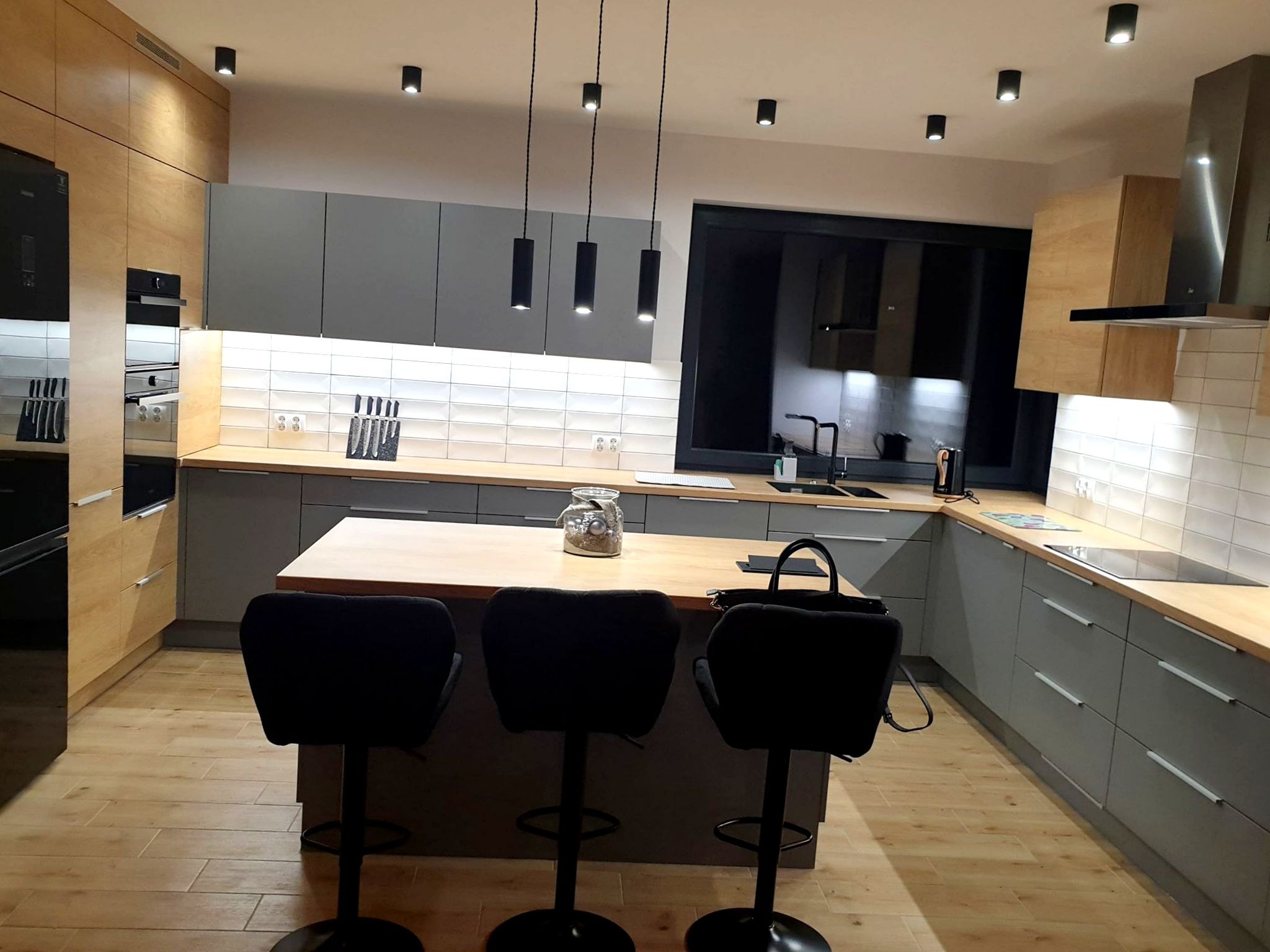 Nowoczesna kuchnia z szarymi szafkami i drewnianymi blatami, oświetlona wiszącymi lampami i punktowymi reflektorami, z wyspą i trzema czarnymi hokerami.