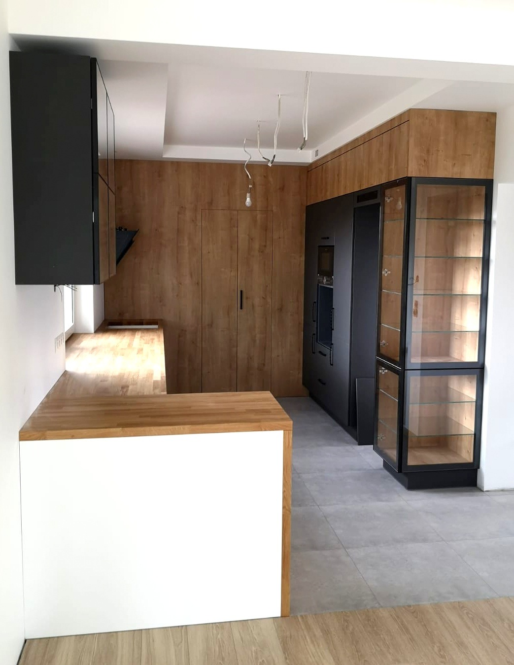 Nowoczesna kuchnia z drewnianymi frontami, czarnymi szafkami i przeszkloną witryną, widoczne niedokończone oświetlenie sufitowe.