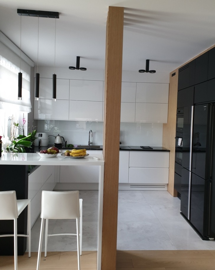 Nowoczesna biała kuchnia z wyspą i drewnianym panelem działowym, oświetlona wiszącymi lampami tubowymi i punktowymi, z owocami na blacie.