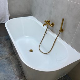 EstetiClean Małgorzata Kufta - Nowoczesna biała wanna wolnostojąca z złotą armaturą na tle szarych płytek i dekoracyjnych płytek podłogowych, z białym ręcznikiem po lewej stronie.