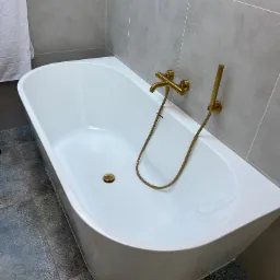 Nowoczesna biała wanna wolnostojąca z złotą armaturą na tle szarych płytek i dekoracyjnych płytek podłogowych, z białym ręcznikiem po lewej stronie.