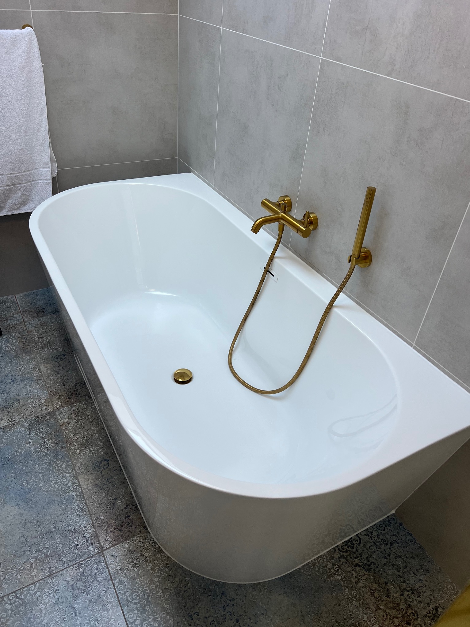 Nowoczesna biała wanna wolnostojąca z złotą armaturą na tle szarych płytek i dekoracyjnych płytek podłogowych, z białym ręcznikiem po lewej stronie.