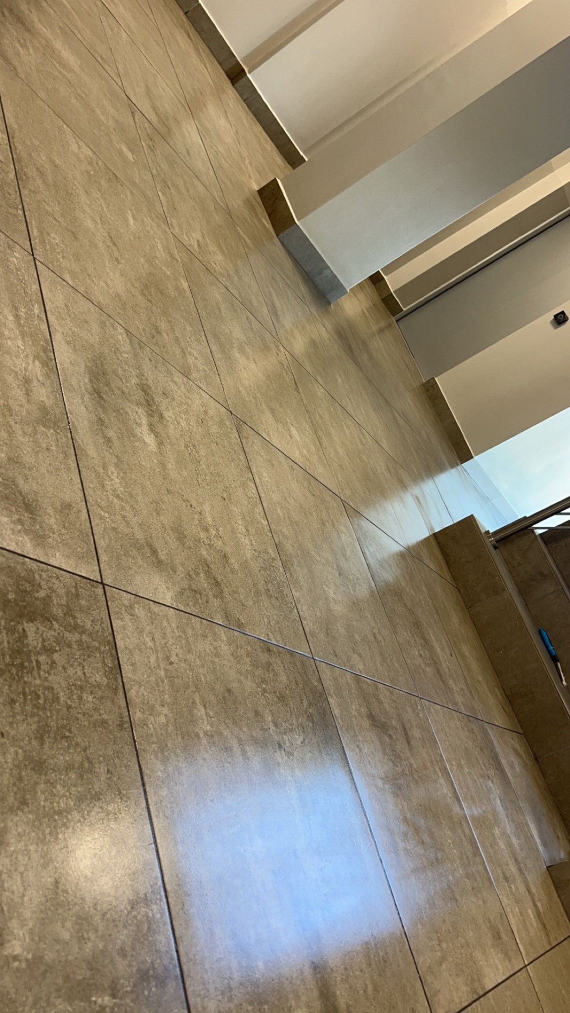 Beżowe, kwadratowe płytki podłogowe z ciemnymi fugami, fragment schodów z metalową barierką w tle, widoczne elementy oświetlenia sufitowego.