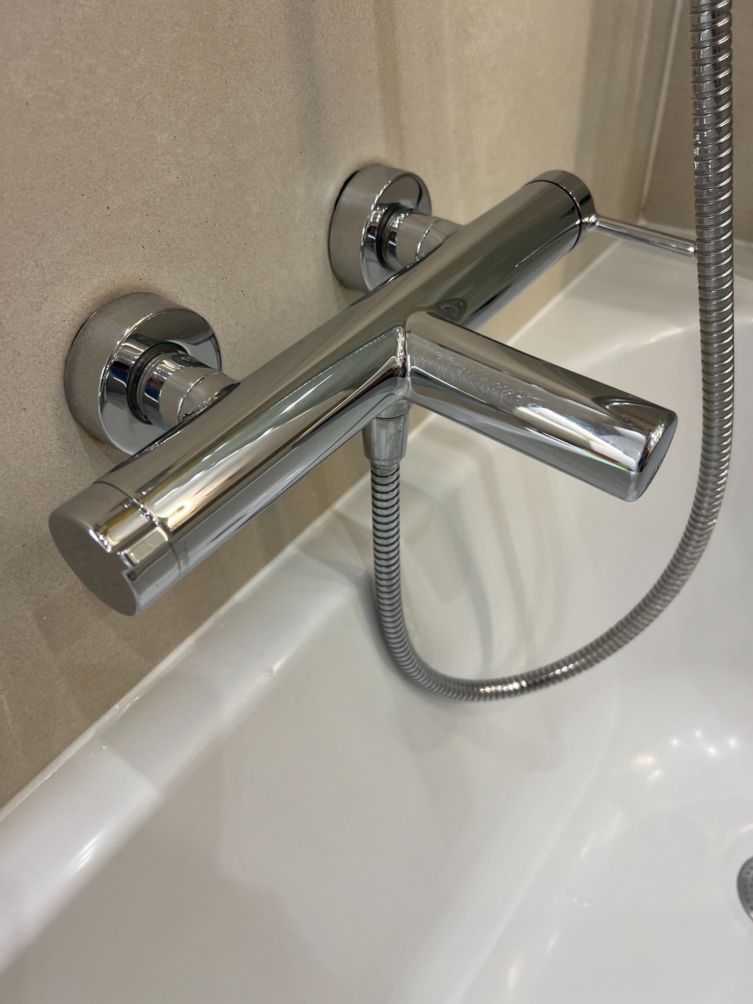 Błyszcząca, chromowana bateria wannowa z wężem prysznicowym zamontowana na beżowej ścianie nad białą wanną.