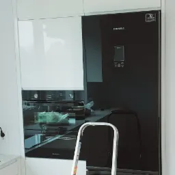 Nowoczesna zabudowa kuchenna w bieli z czarną lodówką Samsung i piekarnikiem, widoczna drabina aluminiowa sugerująca montaż lub wykończenie.