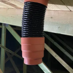 Elastyczny, czarny przewód wentylacyjny z pomarańczowymi łącznikami, zamontowany w drewnianej konstrukcji dachu poddasza.