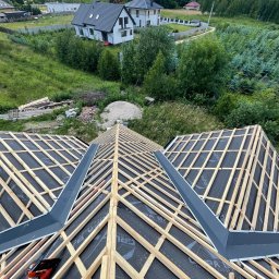 Str. Bud - Konstrukcja dachu z łatami i membraną, widok z góry na tle zieleni i domów. Elementy obróbki blacharskiej w kolorze grafitowym. Na pierwszym planie fragment buta.