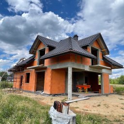 Str. Bud - Dom w trakcie budowy z cegły, z częściowo położonym ciemnym dachem i widocznym rusztowaniem. Na dachu pracuje dekarz. W tle błękitne niebo z chmurami.
