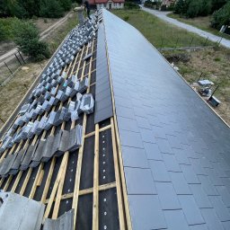 Str. Bud - Układanie grafitowej dachówki na drewnianej konstrukcji dachu domu jednorodzinnego, widok z góry, częściowo pokryty dach, w tle zieleń i zabudowa.