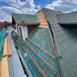 Str. Bud - Dach w trakcie wymiany, widoczne elementy konstrukcyjne i nowe pokrycie. Prace dekarskie na wysokości, rusztowanie z deskami. Błękitne niebo z chmurami w tle.