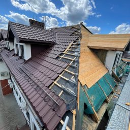 Str. Bud - Częściowo zdjęty brązowy dach z blachy na budynku, widoczne drewniane elementy konstrukcyjne i zabezpieczenia. Błękitne niebo z chmurami w tle.