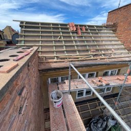Str. Bud - Wymiana dachówki na budynku z cegły, widok z góry na rusztowanie i częściowo ułożone dachówki. Niebo z chmurami w tle.