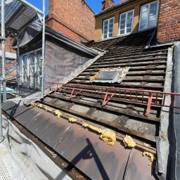 Str. Bud - Dach z częściowo zdjętym pokryciem, widoczne łaty i izolacja piankowa. Na dachu drabina dekarska, w tle budynki z cegły i rusztowanie. Słoneczny dzień.