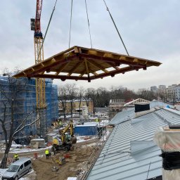 Str. Bud - Montaż drewnianej konstrukcji dachu za pomocą dźwigu na placu budowy. Widoczny fragment dachu z blachy, budynki w tle i pracownicy w odblaskowych kamizelkach.