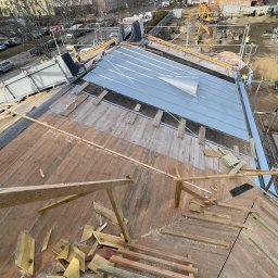Str. Bud - Dach w trakcie krycia blachą trapezową, widoczne drewniane elementy konstrukcji, w tle budynki miejskie i plac budowy z koparkami.