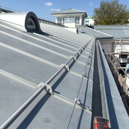 Str. Bud - Szary dach z blachy z systemem odgromowym i lukarną. Widoczny fragment rynny i narzędzie dekarskie. W tle budynek z wieżyczką i drzewo na tle błękitnego nieba.