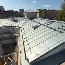 Str. Bud - Metalowy dach z lukarną na tle miejskiej zabudowy w słoneczny dzień. Widoczne elementy konstrukcyjne i rusztowania, sugerujące etap prac budowlanych lub remontowych.
