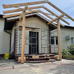 Str. Bud - Konstrukcja drewniana z surowych belek nad wejściem do domu, widoczne rusztowanie, ślady obróbki drewna na kostce brukowej.