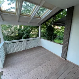 Zadaszony taras z drewnianą podłogą kompozytową, białymi balustradami i oknami dachowymi z widokiem na zieleń.