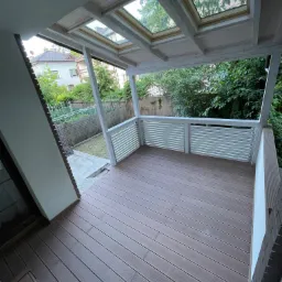 Zadaszony taras z brązową deską kompozytową i białymi balustradami, widok na ogród przez okna dachowe.